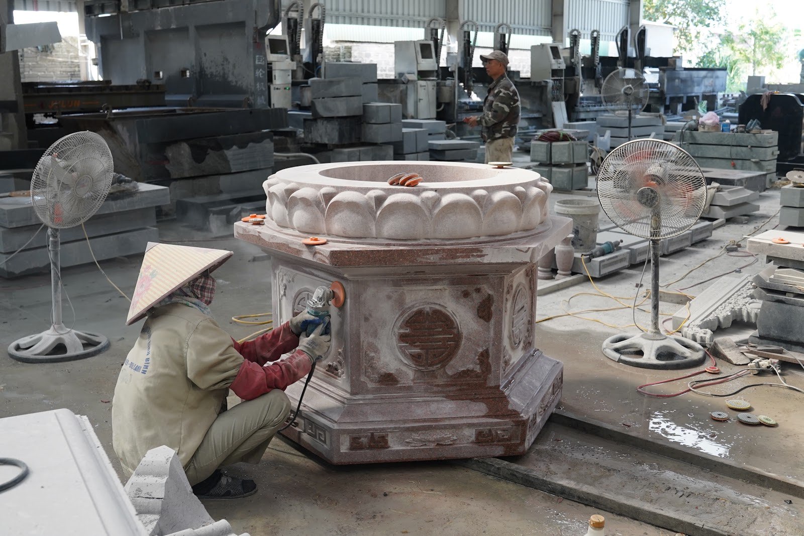 Người thợ đội nón mài phần thân mộ lục giác bằng máy cầm tay trong xưởng, phía sau là các máy CNC